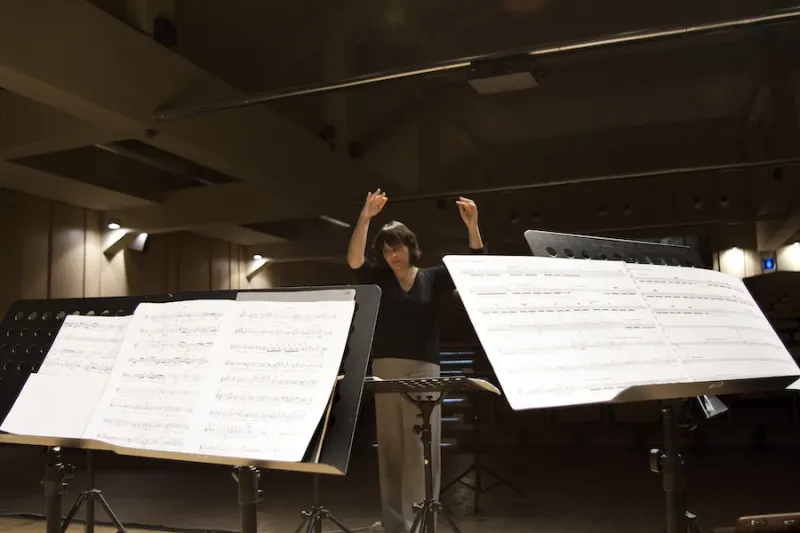 Inquadratura dal basso della direttrice d'orchestra Marilena Solavagione impegnata nella direzione durante una sessione di prove. In primo piano spiccano due grandi leggii con spartiti musicali aperti, mentre lo sfondo mostra la sala illuminata da luci calde.